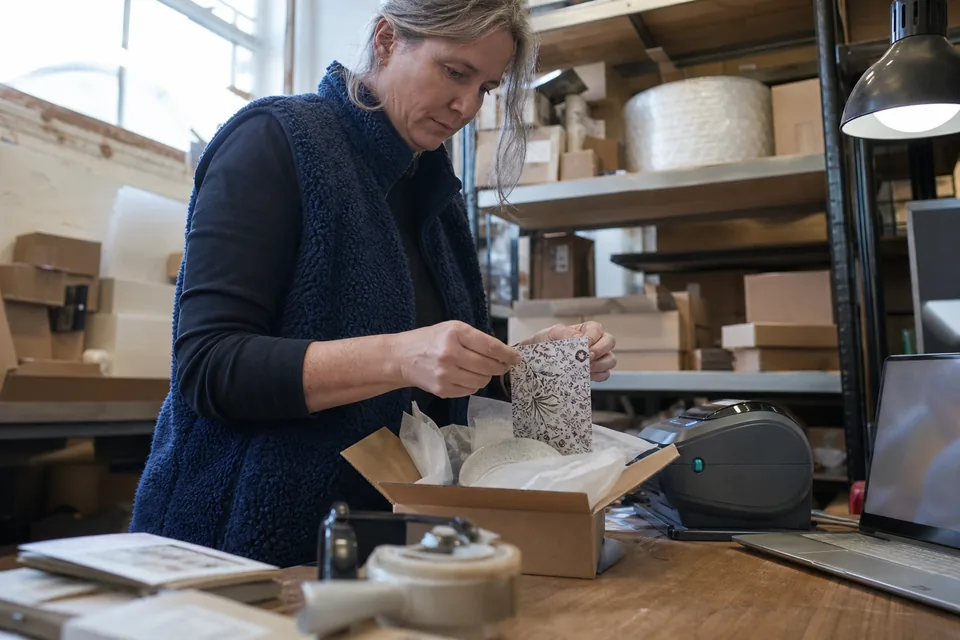 Packer in a small online store stockroom sealing a cardboard parcel and tucking a small printed card inside before closing the lid