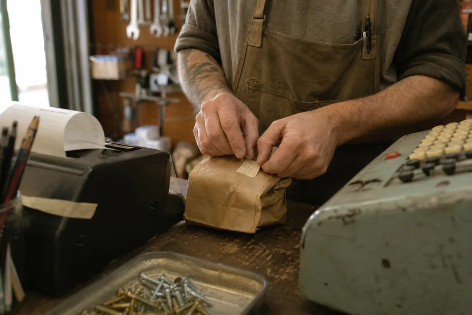 Close detail of a hardware store counter where a clerk sticks a small square code label on a paper bag of fittings next to a receipt roll