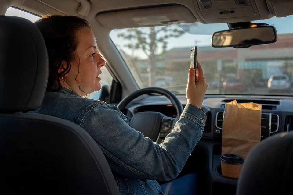 Woman in the driver's seat of a parked car holding her phone up in front of her to record a short video of herself, late afternoon light