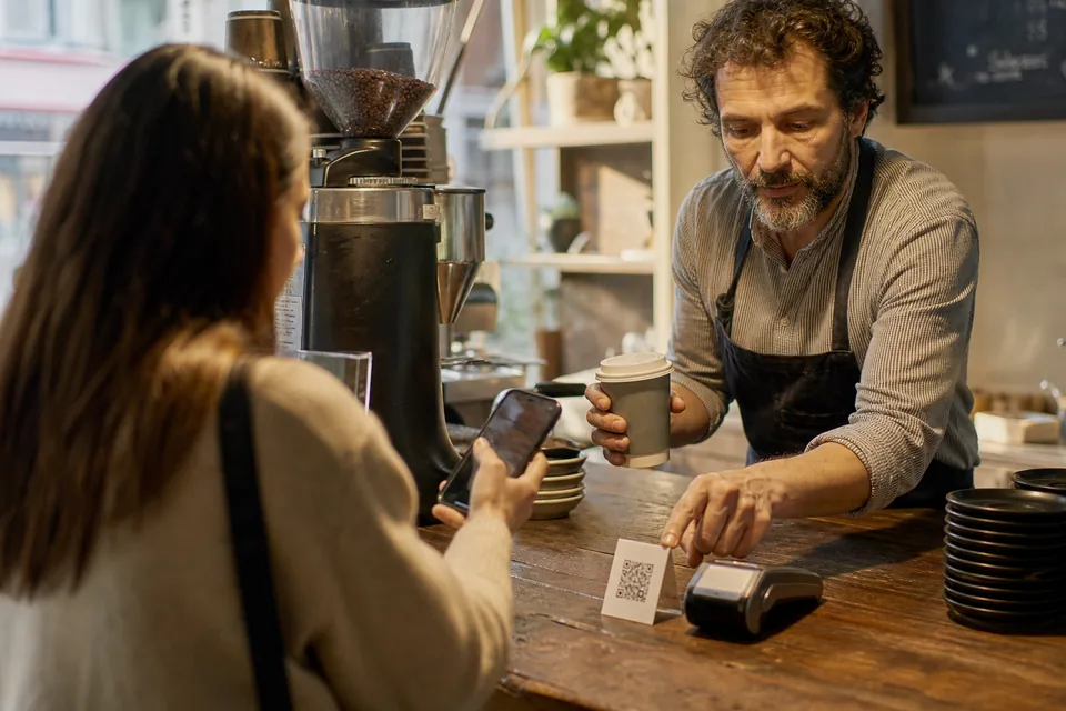 Cafe owner at the espresso counter pointing a regular customer to a small standing card with a square code next to the card reader while handing back her cup